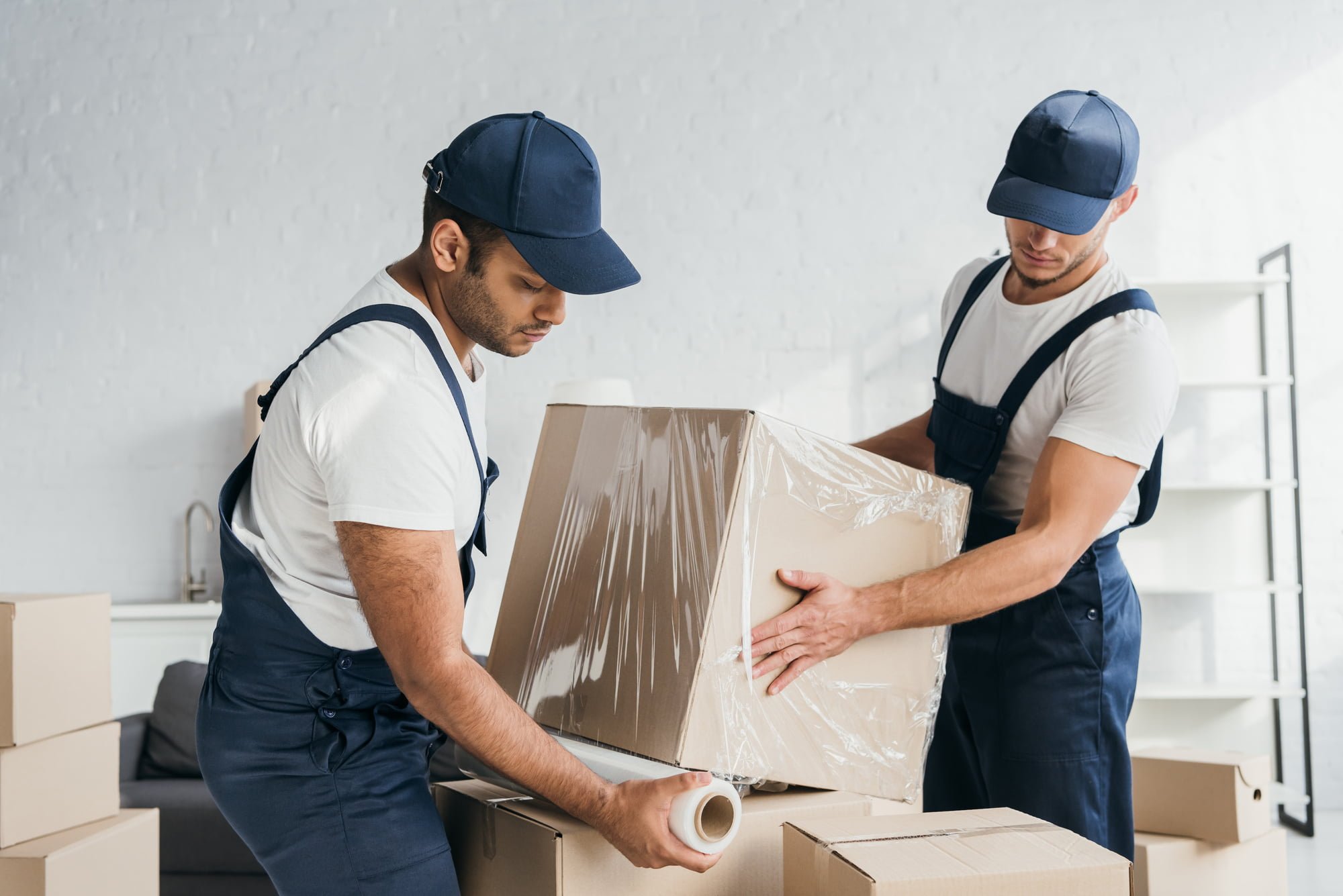 multiethnic movers in overalls wrapping box with stretch film